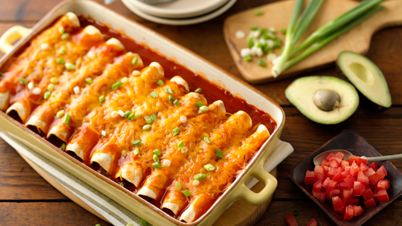 A baking dish filled with cheese-topped Easy Ground Beef Enchiladas, garnished with sliced green onions, surrounded by diced tomatoes, avocado, extra green onions, and plates, all set on a wooden table.