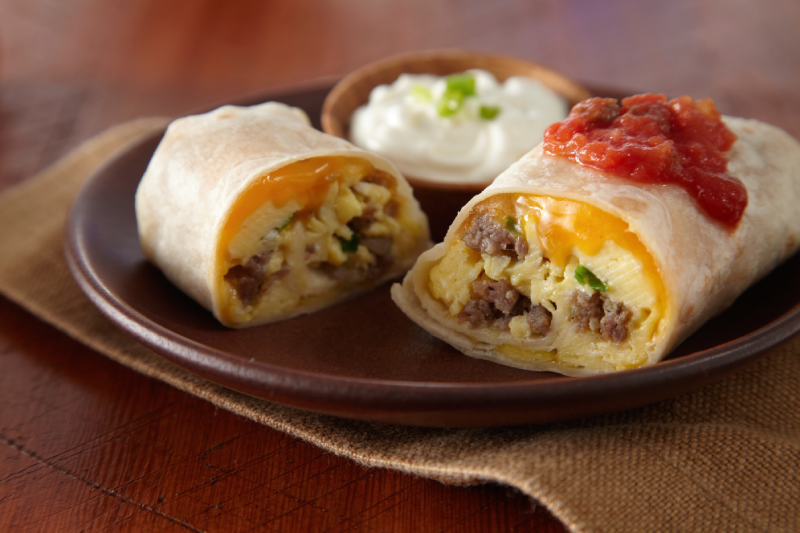 An egg, sausage and cheese breakfast burrito cut in half with salsa and sour cream on a brown plate.